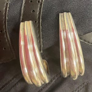 May include: A pair of silver-toned earrings with a ribbed, curved design. The earrings are set against a black background, with a leather strap visible in the upper left corner. The earrings have a polished finish.