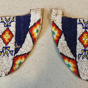 May include: A pair of U-shaped beaded patches featuring intricate geometric patterns in dark blue, white, red, yellow, and green glass beads. The design includes a central white vertical band with a red diamond and symmetrical blue accents.