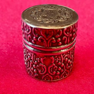 May include: A small cylindrical metal container with a removable lid. The exterior features a deep red enamel finish with raised silver-toned scrollwork and floral patterns. The top of the lid displays a detailed embossed circular floral design.