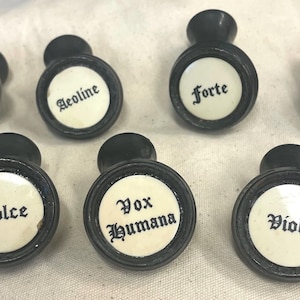 May include: A collection of seven antique black and white drawer pulls. Each pull has a circular white center with a black border and a black knob. The centers are labeled with musical terms: Melodia, Aeoline, Forte, Diapason, Dolce, Vox Humana, and Viola.