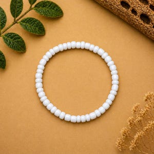 May include: A white beaded bracelet, circular in shape, rests on a tan surface. The bracelet is composed of small, rounded white beads. Green leaves and a piece of wood are in the background, adding a natural aesthetic. This bracelet is a simple, elegant accessory.