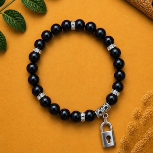 May include: A beaded bracelet featuring smooth, round, glossy black beads interspersed with small, silver-toned spacers encrusted with clear crystals. A silver-toned metal padlock charm hangs from the bracelet, featuring a keyhole cutout.