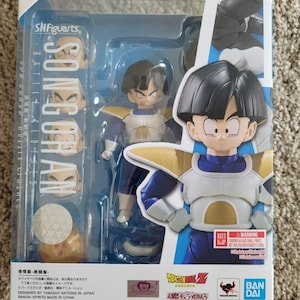 May include: A plastic box containing a Son Gohan action figure from the Dragon Ball Z anime series. The figure is in a blue and yellow outfit with a yellow belt and a yellow symbol on the chest. The box also includes a stand and a few extra parts for the figure. The box has a warning label for children under 3 years old.