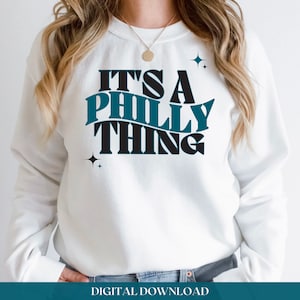 Könnte beinhalten: Weißer Pullover mit dem Text "It's a Philly Thing" in schwarzer und blaugrüner Schrift mit zwei kleinen Sternen auf jeder Seite des Textes.