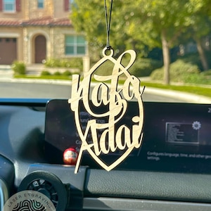 May include: A wooden car charm with the names "Hafa Adai" engraved in a cursive font. The charm is hanging from a black string and is in focus against a blurred background of a house and trees.