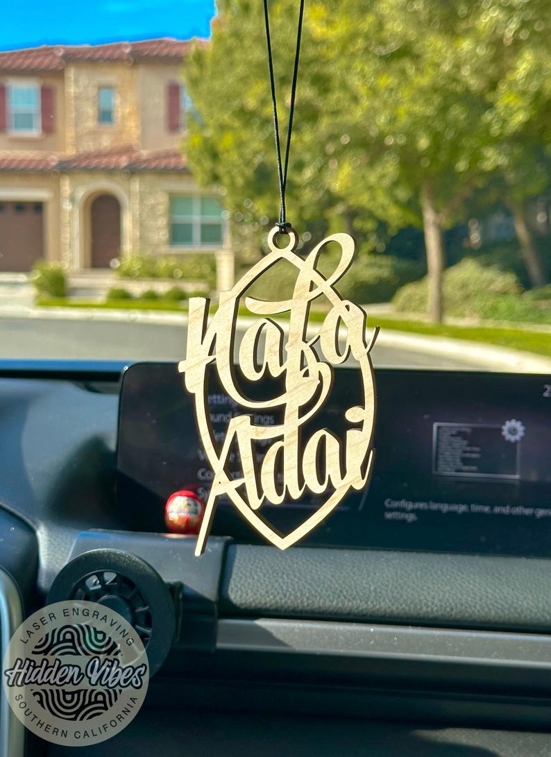 May include: A wooden car charm with the names "Hafa Adai" engraved in a cursive font. The charm is hanging from a black string and is in focus against a blurred background of a house and trees.