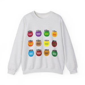 May include: A light gray sweatshirt featuring a pattern of colorful, stylized cans. Each can has a different color and design, with the text "SweetBalls" visible on each. The cans are arranged in a grid pattern across the front of the sweatshirt.