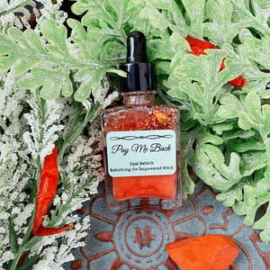 May include: A clear glass bottle with a dropper top filled with a red liquid. The bottle has a white label with the text "Pay Me Back Opal Rebirth Rebirthing the Empowered Witch".