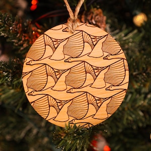 May include: A round wooden Christmas ornament with a laser-etched design of a flock of geese flying in formation. The ornament is a light brown color and has a white string for hanging.