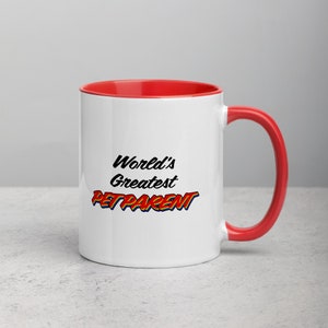 Könnte beinhalten: Weiße Keramiktasse mit rotem Rand und Henkel. Die Tasse trägt den Text "World's Greatest Pet Parent" in schwarzer, roter und blauer Schrift.