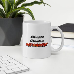 Pode incluir: Caneca de cerâmica branca com o texto "World's Greatest PETPARENT" em preto, vermelho e azul. A caneca tem uma alça curva. Uma planta e livros são visíveis no fundo, sobre uma superfície branca.