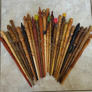May include: A collection of twenty-five handcrafted wooden wands arranged in a fan shape. Each wand features unique carvings, varying wood tones from light to dark, and decorative finials including spheres, points, and colored resin accents.