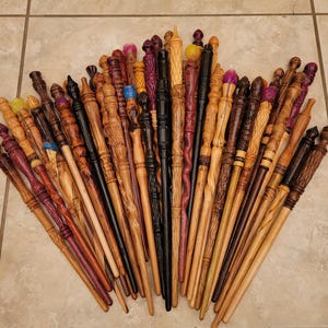 May include: A collection of handcrafted wooden wands in various colors and designs. The wands vary in length and feature intricate carvings, painted details, and unique handle shapes. Some wands have colorful crystal or glass accents.