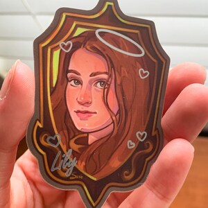 Op de afbeelding: Een sticker met een portret van een vrouw met bruin haar, die een halo draagt. Het portret is ingelijst in een gouden lijst met een bruine achtergrond. De sticker heeft een wit hartontwerp en het woord "Lily" in zilveren letters.
