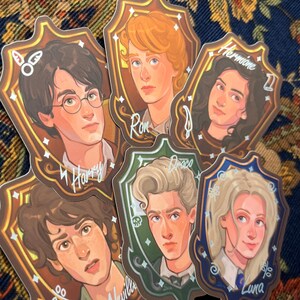 Może przedstawiać: Sześć ilustrowanych naklejek z postaciami z serii Harry Potter. Każda naklejka to portret postaci w ramce w kształcie tarczy z imieniem napisanym poniżej. Postaciami są Harry, Ron, Hermiona, Draco, Luna i Neville.