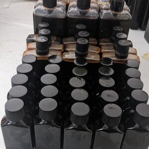 Może przedstawiać: Kolekcja małych, ciemnobrązowych butelek szklanych z czarnymi zakrętkami. Butelki są ułożone w rzędach i kolumnach na białym tle.