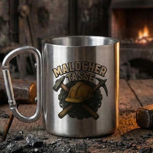 Peut inclure: Un mug en acier inoxydable avec une anse mousqueton, orné d'un graphisme représentant un casque de chantier jaune et deux pioches croisées, avec le texte Malocher Tasse imprimé en lettres noires et dorées sur le devant du récipient cylindrique.