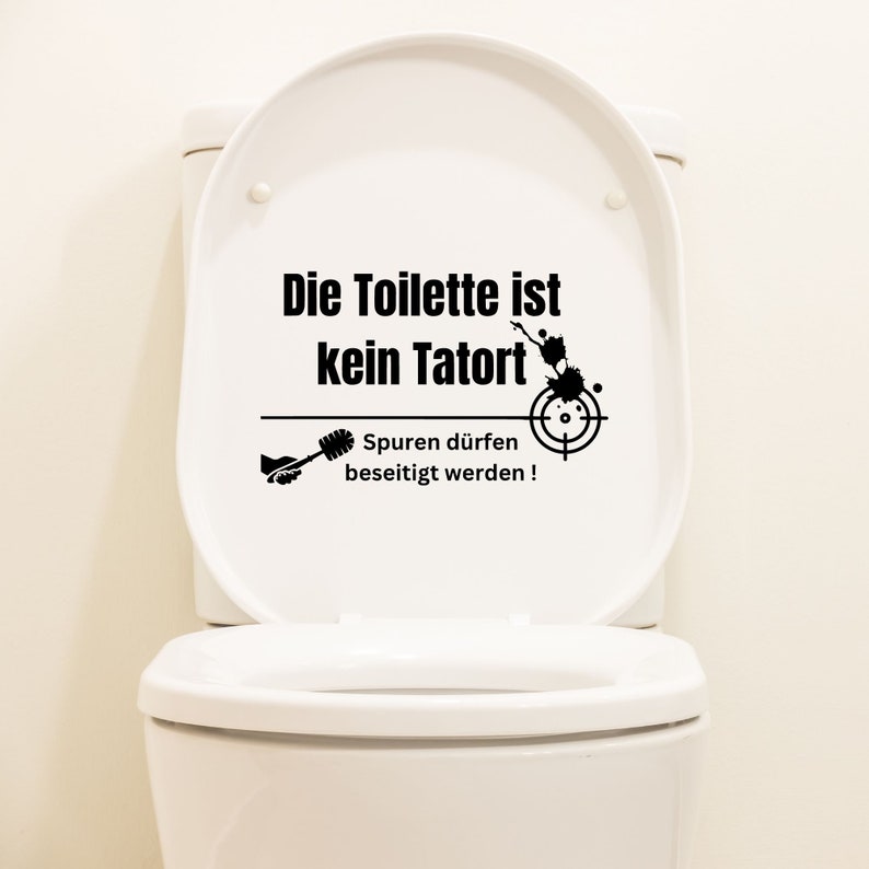 K&ouml;nnte beinhalten: Eine wei&szlig;e Toiletten-Sch&uuml;ssel mit einem schwarzen Aufkleber, auf dem "Die Toilette ist kein Tatort" und "Spuren d&uuml;rfen beseitigt werden!" steht, sowie ein Ziel und eine Toilettenb&uuml;rste.
