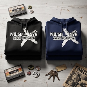 Könnte beinhalten: Ein schwarzer Kapuzenpullover und ein dunkelblauer Kapuzenpullover, jeweils mit einem weißen Grafikdruck mit dem Text NO.50 - 1976 BADASS GENERATION und einem großen X. Die Pullover haben passende Kordelzüge und eine Vordertasche, neben Kassetten.