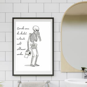 May include: A framed black and white print featuring a skeleton holding a roll of toilet paper. The skeleton is depicted in a thoughtful pose. The print includes German text. The print is hanging in a bathroom.