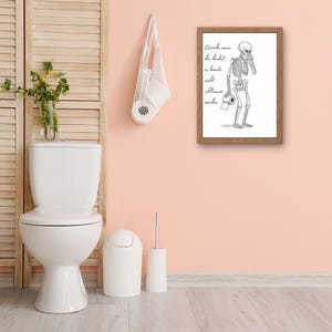 May include: A framed black and white illustration of a skeleton holding toilet paper, with cursive text. The bathroom scene includes a white toilet, bin, and toilet brush, set against a pink wall and wooden floor.