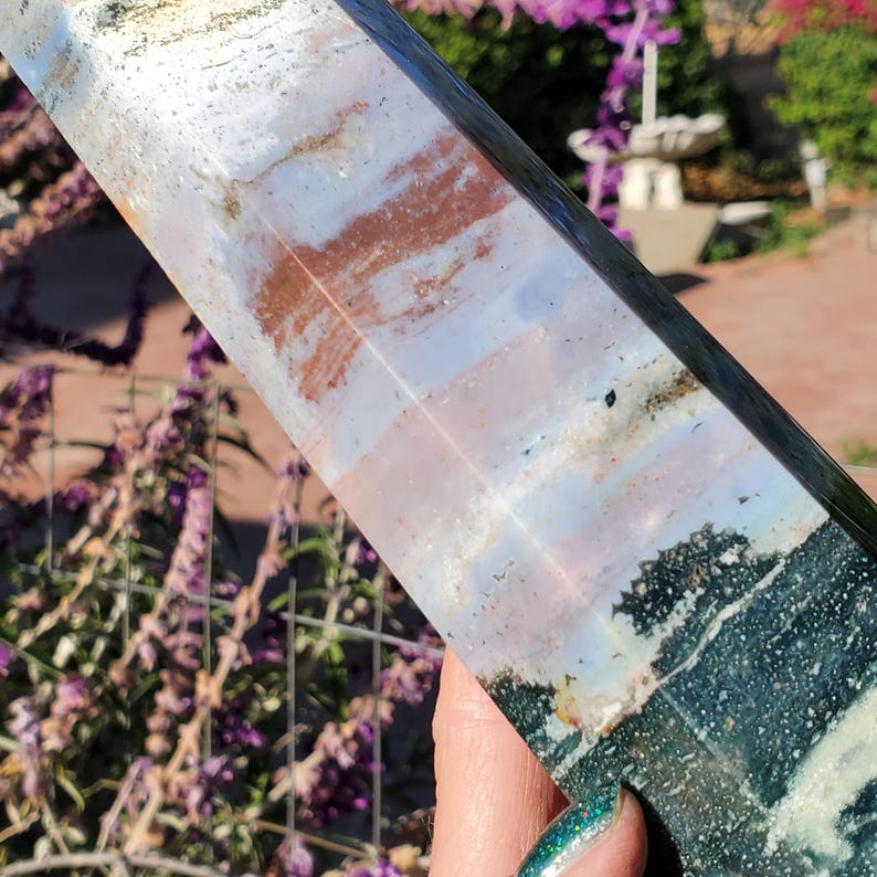 May include: A polished stone tower featuring a multi-colored surface with bands of white, soft pink, and deep rust-orange, transitioning into a dark, textured forest green base with intricate mineral inclusions and a smooth, reflective finish.