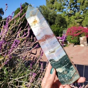 May include: A polished, multi-colored crystal tower featuring a hexagonal point. The stone displays a layered pattern with bands of deep forest green, earthy brown, soft lavender, and creamy white, with a small golden-brown inclusion near the top.