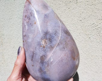 May include: A large, polished stone with a smooth, rounded shape. The stone features a blend of lavender, gray, and brown hues, with subtle veining and a glossy finish. The overall form is reminiscent of a teardrop or egg shape.