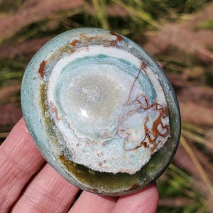 May include: A polished, oval-shaped stone with a swirling pattern of colors. The stone features concentric rings of light blue, white, brown, and green hues. The stone is held in a hand, with a blurred background of green grass.