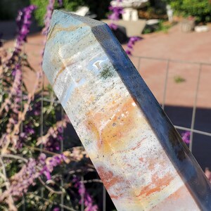 May include: A polished stone tower with a hexagonal point, featuring a marbled pattern of white, gray, tan, and burnt orange hues. The surface displays natural inclusions and translucent bands that catch the light, creating a smooth, multifaceted appearance.