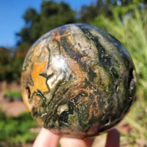 May include: A polished stone sphere with a swirling pattern of green, brown, and yellow hues. The orb is held in a hand, showcasing its natural colors and textures. The sphere's surface has a smooth, glossy finish.