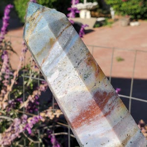 May include: A polished, multi-faceted crystal tower featuring a blend of pale blue, white, and earthy brown bands. The stone displays natural inclusions, translucent layers, and a pointed top, showcasing a smooth, reflective surface with varied mineral patterns.