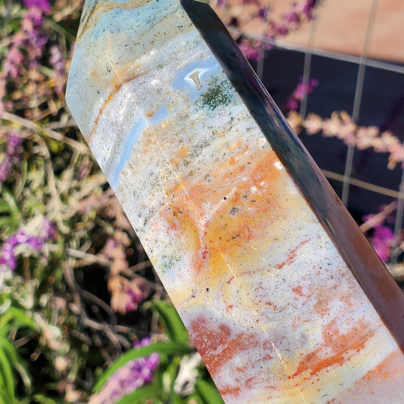 May include: A polished stone tower with a hexagonal shape, featuring a blend of translucent light blue, creamy white, and warm orange-brown bands. The surface displays natural mineral inclusions, speckles, and smooth, reflective facets.