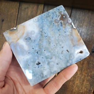 Könnte beinhalten: Ein quadratischer, polierter Stein mit einem einzigartigen Muster aus blauen, weißen und beigen Wirbeln und Flecken. Der Stein hat eine glänzende Oberfläche und wird in einer Hand gehalten. Die Oberfläche des Steins hat ein natürliches, organisches Aussehen.