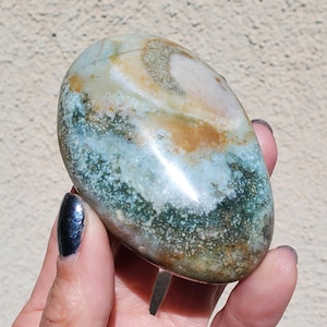 May include: A polished, oval-shaped stone with a blend of colors. The stone features areas of light blue, green, brown, and cream, creating a marbled effect. The surface has a smooth, glossy finish, highlighting the natural patterns.