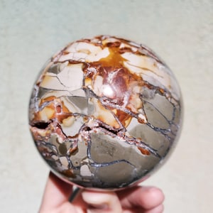 May include: A spherical decorative stone with a cracked surface. The stone features a mix of colors, including beige, brown, and gray, with dark gray veins. The sphere has a rough texture and a natural, earthy appearance.