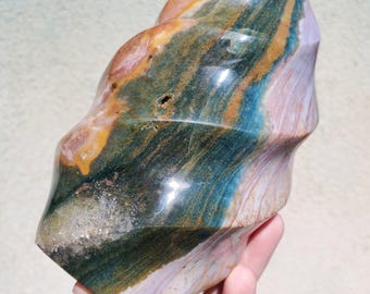 May include: A polished, freeform stone sculpture with a swirling pattern of colors. The stone features bands of green, gold, pink, and blue. The sculpture has a smooth, glossy finish and is held in a hand.