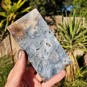 Könnte beinhalten: Eine rechteckige Steinplatte mit einem einzigartigen Muster aus weißen, grauen, braunen und schwarzen Wirbeln und Flecken. Die polierte Oberfläche wird in einer Hand gehalten und zeigt die natürlichen Variationen in Textur und Farbe des Steins.