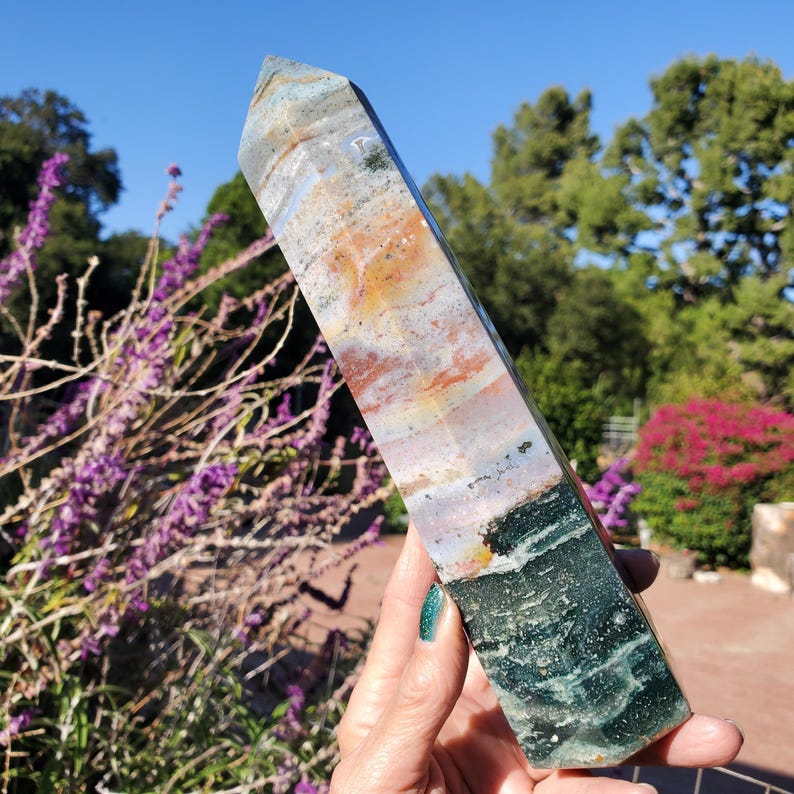 May include: A polished, hexagonal crystal tower featuring a multi-colored surface with bands of deep forest green, earthy brown, soft orange, and translucent grey. The stone has a smooth, faceted finish that tapers to a point at the top.