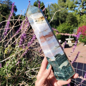 May include: A polished, hexagonal crystal tower featuring a multi-colored surface with distinct bands of deep forest green, soft lavender, muted blue, and earthy brown tones. The stone has a smooth, faceted finish that tapers to a point.