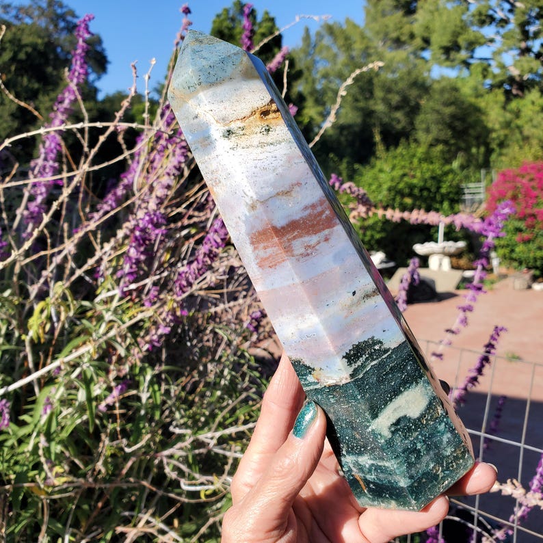 May include: A polished, hexagonal crystal tower featuring a multi-colored surface with distinct bands of deep forest green, soft lavender, muted blue, and earthy brown tones. The stone has a smooth, faceted finish that tapers to a point.