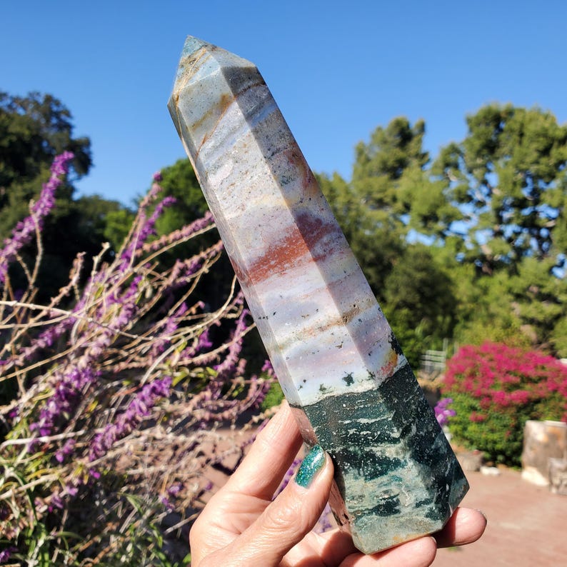 May include: A tall, hexagonal crystal tower featuring a distinct color gradient. The top section displays bands of pale blue, white, and reddish-brown, while the bottom portion transitions into a deep, solid forest green with dark mineral inclusions.