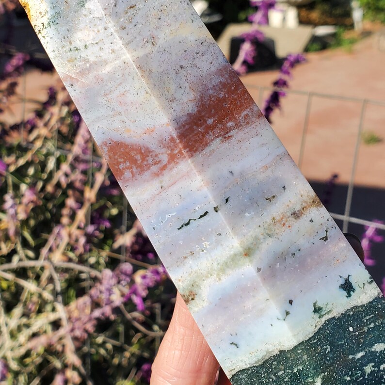 May include: A polished stone tower with a multi-faceted surface featuring horizontal bands of white, translucent gray, rust orange, and deep forest green. The mineral specimen displays natural inclusions and varied color gradients throughout its length.