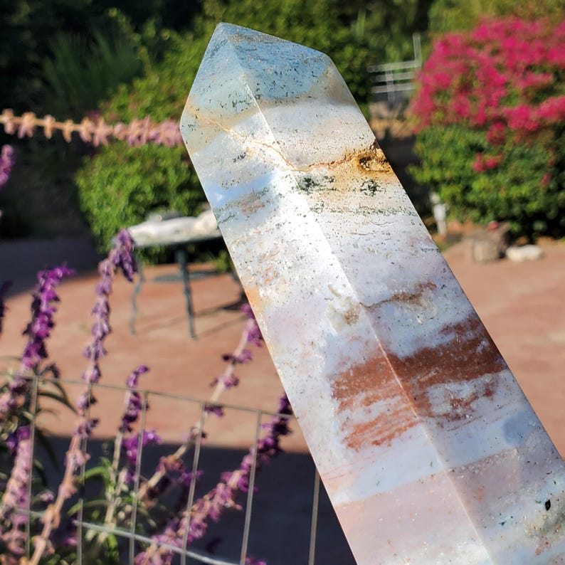 May include: A polished, hexagonal crystal tower featuring a blend of translucent light blue, milky white, and earthy brown bands. The stone displays natural inclusions, dark mineral flecks, and a smooth, faceted surface that tapers to a point.