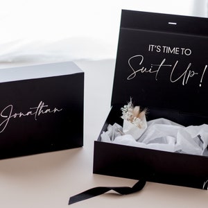 May include: Black gift box with the text "It's time to Suit Up!" on the lid. The box is open and contains a small bouquet of dried flowers. The box is personalised with the name "Jonathan".