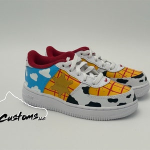 Op de afbeelding: Witte leren sneakers met een aangepast handgeschilderd ontwerp met Woody uit Toy Story. Het ontwerp bevat een blauwe lucht met wolken, een gele sheriffster en een rood-geel ruitpatroon.