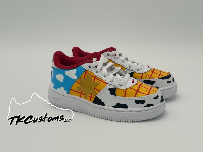 Op de afbeelding: Witte leren sneakers met een aangepast handgeschilderd ontwerp met Woody uit Toy Story. Het ontwerp bevat een blauwe lucht met wolken, een gele sheriffster en een rood-geel ruitpatroon.