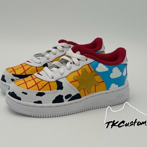 Op de afbeelding: Witte lage sneakers met een aangepast ontwerp met een geel en rood geruit patroon, een zwart-witte koe-print, een gele ster en een blauwe lucht met witte wolken. De sneakers hebben een rode tong en een witte zool.