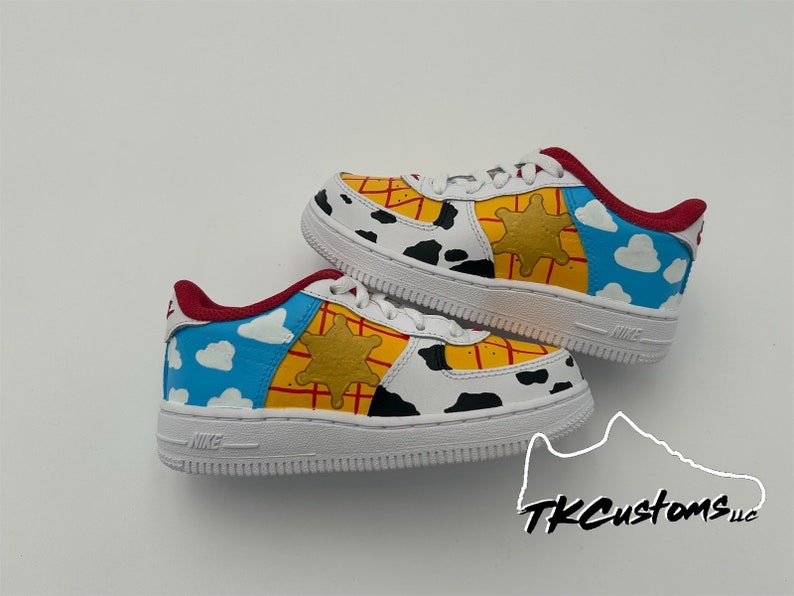 Op de afbeelding: Witte sneakers met een aangepast ontwerp met een Toy Story-thema. De schoenen hebben een blauwe lucht met witte wolken, een gele sheriffster en een rood-geel ruitpatroon.