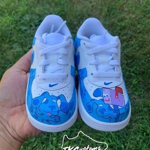 Op de afbeelding: Paar op maat gemaakte witte en blauwe sneakers met een cartoon hond ontwerp. De schoenen hebben witte veters, een blauwe Nike swoosh en een cartoon hond karakter op de teen. Op een groene achtergrond.